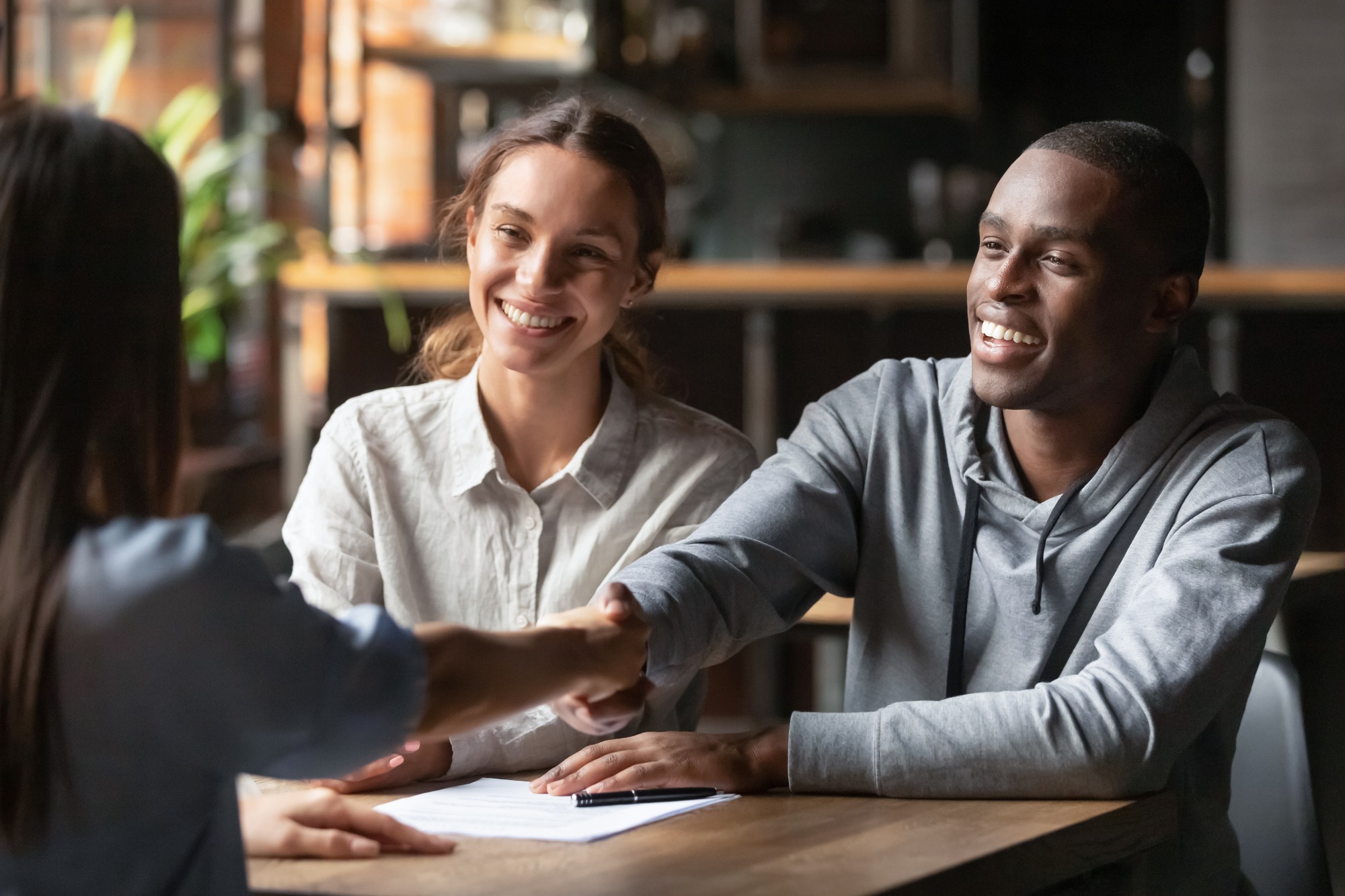 Happy Young Interracial Couple Shake Hand of Bank Manager Broker Buy Insurance Services Take Mortgage Loan, Mixed Ethnicity Customers Handshake Agent Make Agreement Financial Business Deal at Meeting.